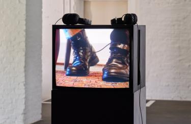 Ein schwarzer, quadratischer Röhrenfernseher steht auf einem dunkelgrauen Sockel im Ausstellungsraum der Filmwerkstatt. Auf dem Monitor liegen zwei kabelverbundene Kopfhörer. Der Fernseher zeigt ein schwarzes Paar blankpolierter Lederstiefel auf einem ornamentalen turkmenischen Buchara-Teppich.