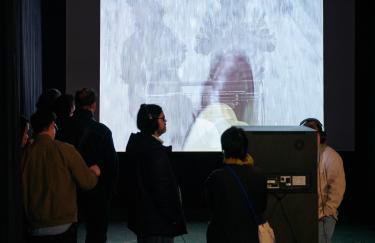 Die zweikanalige Videoinstallation „Auf Schritt und Tritt“ von Marcel Odenbach wird von Besuchenden angeschaut. Am Ende des Raumes hängt eine große Projektionswand, die eine Szene mit zwei übereinandergelegten Bildmotiven zeigt: ein Lederschuh des Künstlers und spielende Kinder. In der Mitte des abgedunkelten Raumes steht ein schwarzer CRT MonitorSockel, der zur Leinwand hin ausgerichtet ist. Zwei Besuchende stehen neben diesem Fernseher und tragen damit verbunde Kopfhörer.