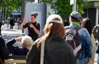 Roman Poryadin steht auf dem Carlsplatz in Düsseldorf vor einer Führungsgruppe und erzählt ihnen, dass hier ein Bildschirm zu sehen ist, der Seo Hye Lees „Ohrenbetäubende Stille“ im öffentlichen Raum abspielt.