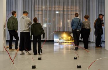Ein Blick in die Ausstellung im Storage Museum. Am Ende des Raums steht ein großer Fernseher auf dem Boden und zeigt Seo Hye Lees „Ohrenbetäubende Stille“. Davor stehen Besuchende, die sich das Werk anschauen.