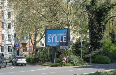 Eine Düsseldorfer Straßenkreuzung zur Mittagszeit. Die große Mittelinsel wird vollständig vom Grün verschiedener Bäume, Sträucher und Gräser eingenommen. Davor steht eine digitale Werbetafel, auf der Seo Hye Lees „Ohrenbetäubende Stille“ zu sehen ist. Die Ampel für Linksabbieger hat gerade auf grün geschaltet und ein paar Autos fahren nun los. Der andere Fahrstreifen hat rot und ist momentan komplett leer. Im Hintergrund sind Reihenhäuser zu sehen.