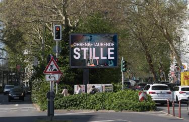 Eine mehrspurige Straßenallee bei hellem Tageslicht. Links kommen uns Autos entgegen, rechts fahren sie davon. Auf dem begrünten Mittelstreifen steht eine digitale Werbetafel, auf der Seo Hye Lees „Ohrenbetäubende Stille“ zu sehen ist. Davor befinden sich Passanten mit Kinderwagen sowie ein Baustellenschild.