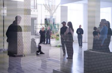 Ein Blick durch ein Fenster in das Storage Museum, wo mehrere Menschen durch die Ausstellung gehen und sich zum Teil darüber austauschen. Im Fensterglas spiegelt sich der Innenhof, wo ebenfalls Besuchende stehen und sich unterhalten.